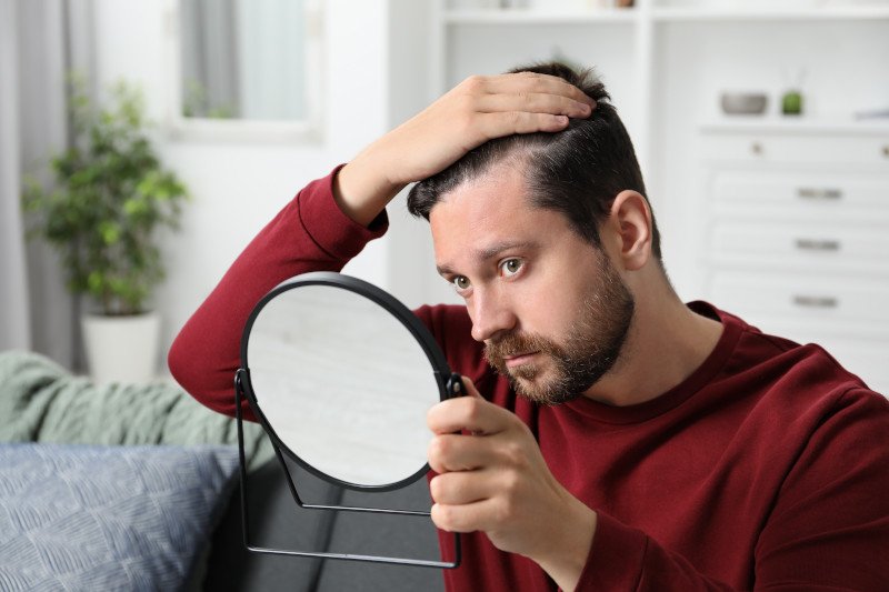 Ein Mann schaut seine Haare im Spiegel an und spielt mit dem Gedanke einer Haartransplantation